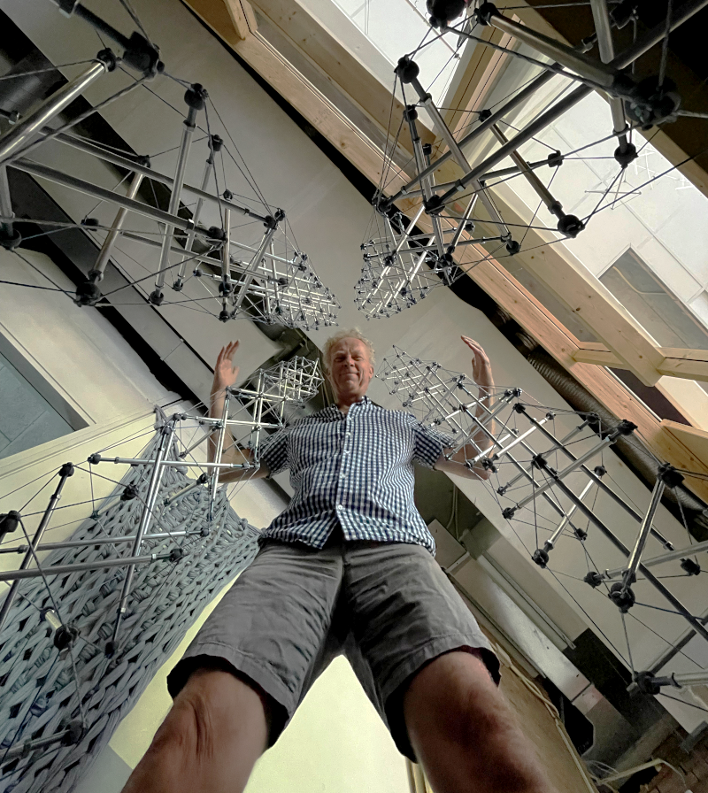 Gerald standing in the middle of three tensegrity builds, camera angled from the floor upward.